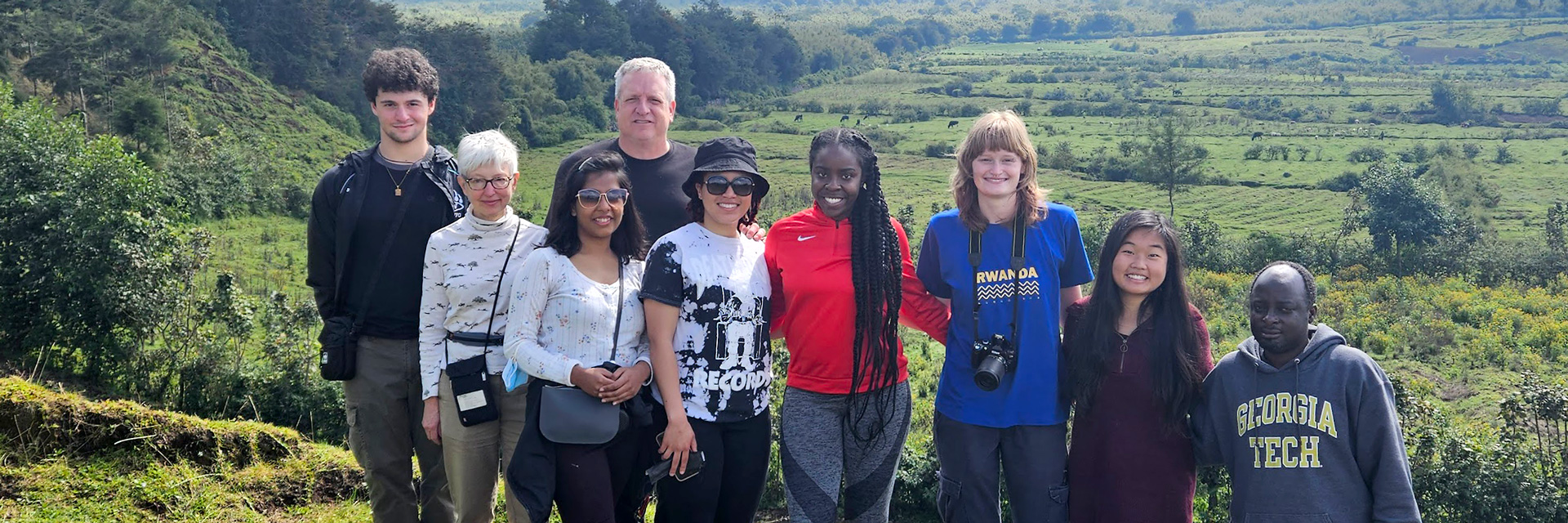 Group photo of six Georgia Tech students and two faculty with guide in Rwanda, with farmland in the background 