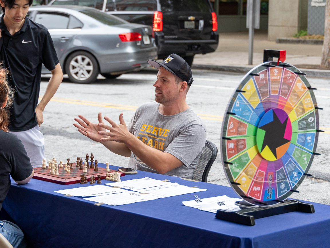 Alex Kozela seated at a table in a parking space, talking to pedestrians. A prize wheel labeled with the UN's Sustainable Development Goals is on the table.