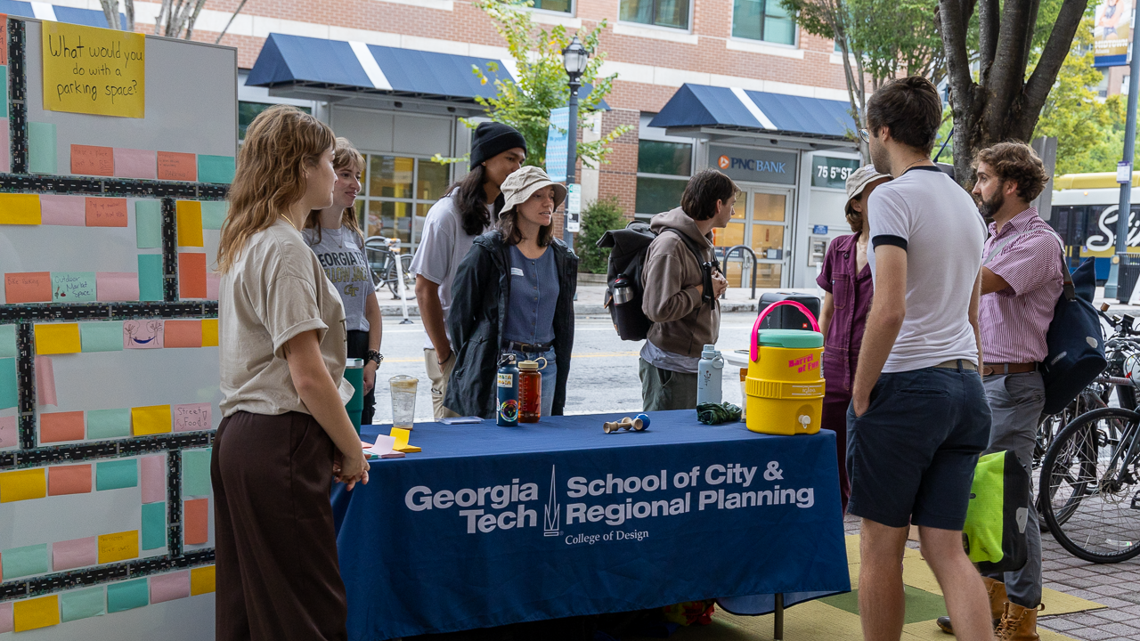 Student Planning Association members at a table outdoors talking to pedestrians. An idea board for parking space reuse stands on the left side. The tablecloth has the logo of the Georgia Tech School of City and Regional Planning.